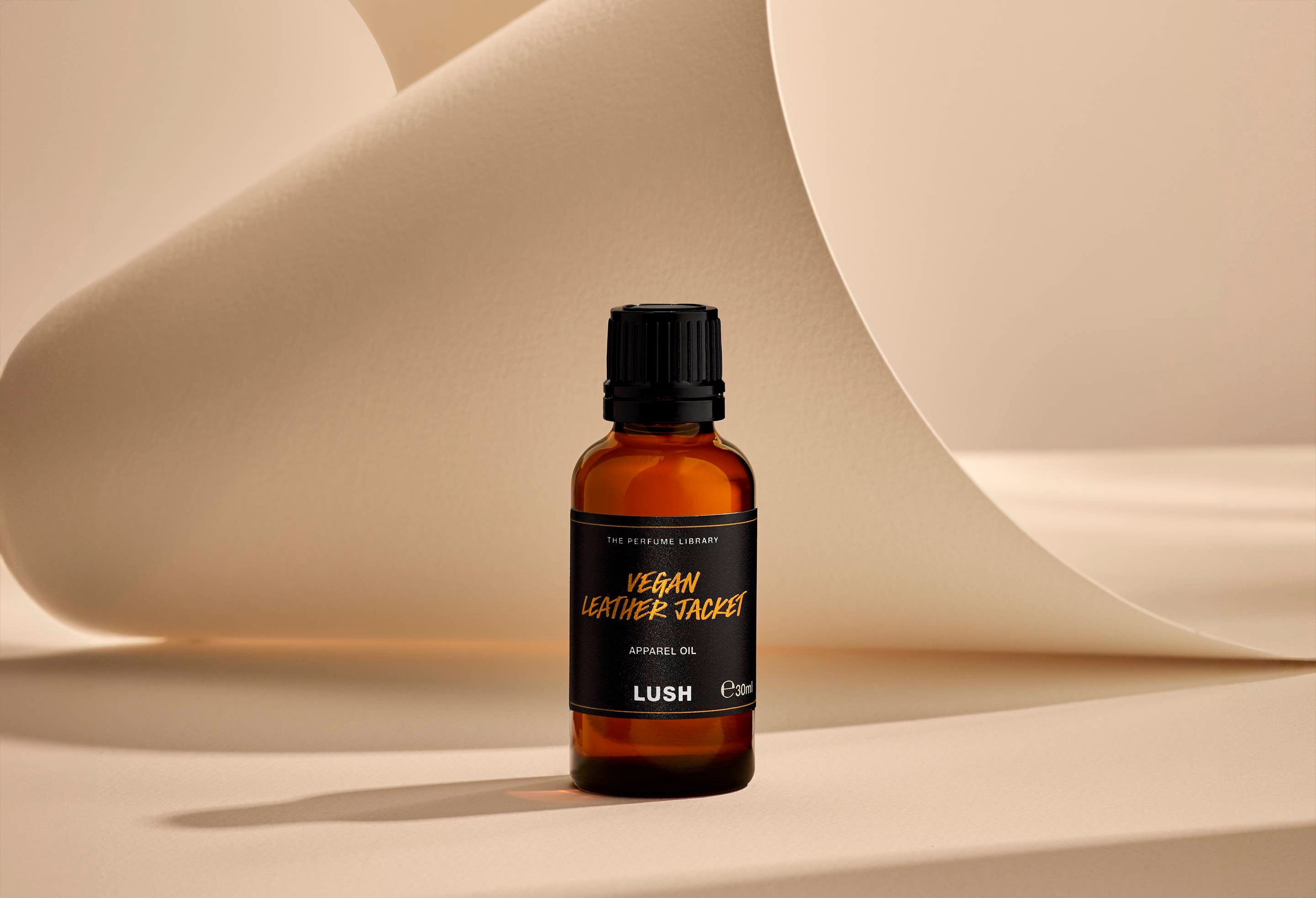 The image shows the Vegan Leather Jacket, brown glass, apparel oil bottle. It is sitting on a soft, beige background with curved, cream paper creating soft, gliding shapes behind the product.