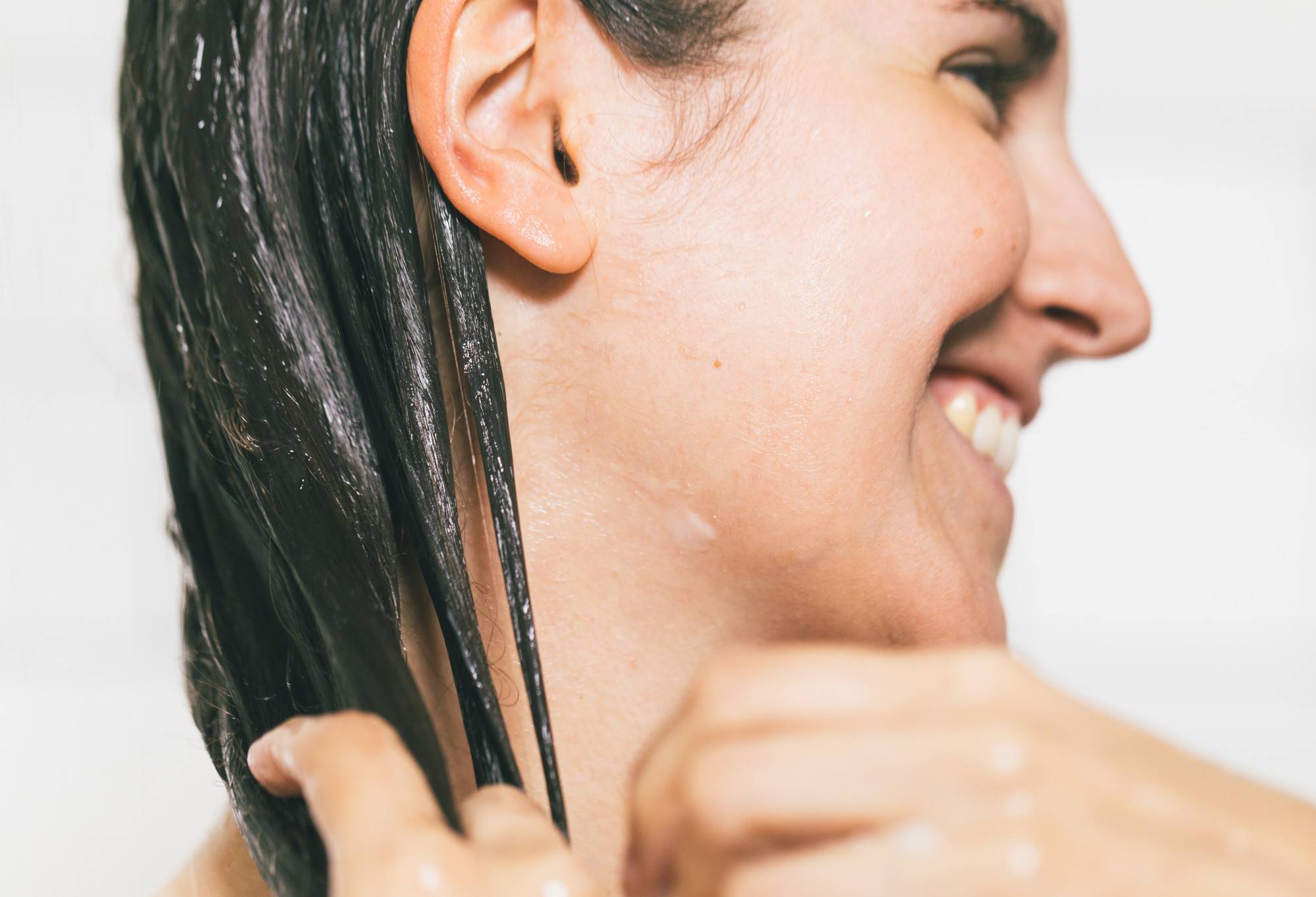 A beaming person with long, dark, conditioner coated hair hair, runs their hands through their lengths, detangling the hair.
