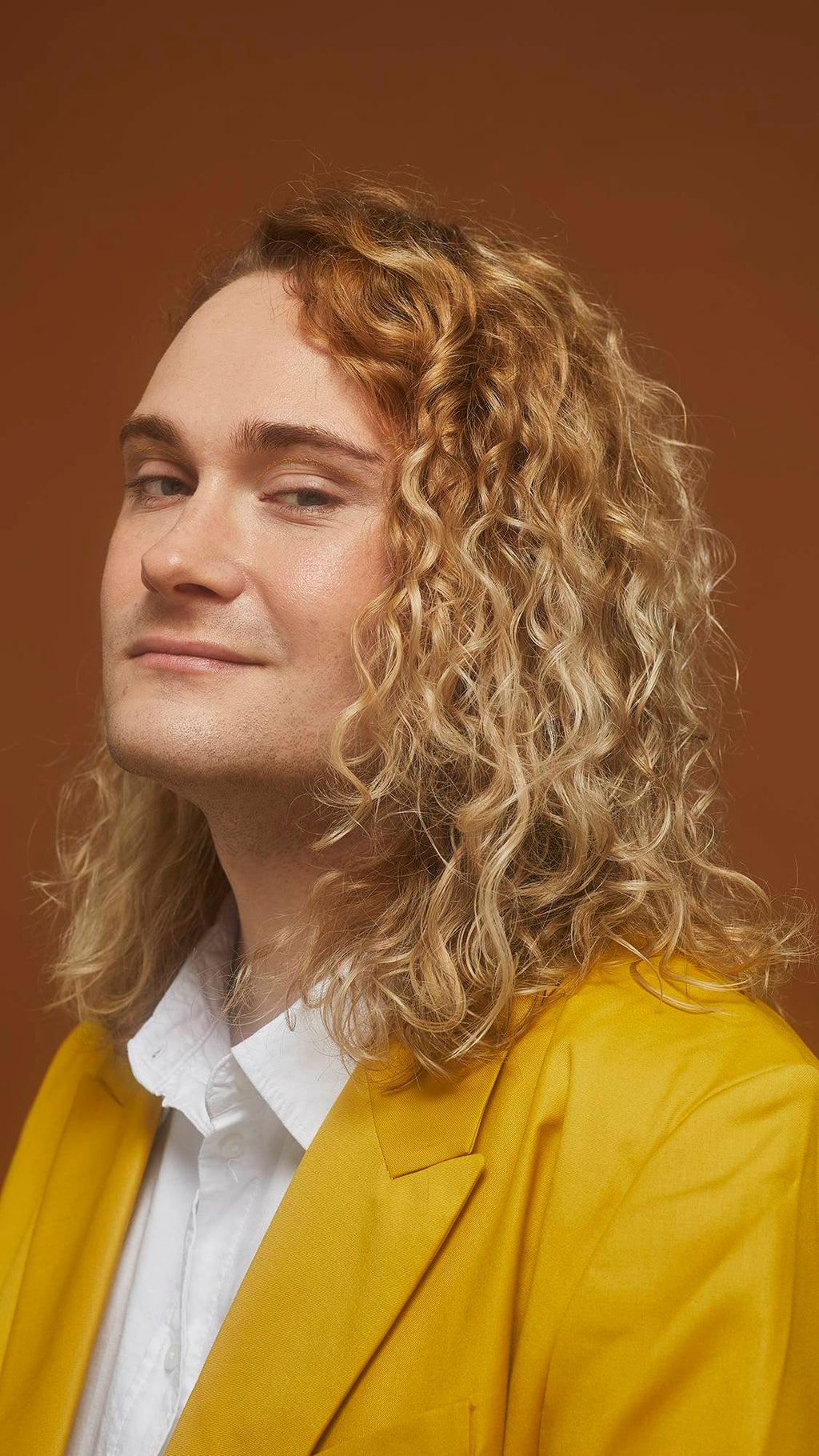 Model smiles and wears a bright yellow jacket showing defined, golden, curled locks on an earth-toned background.