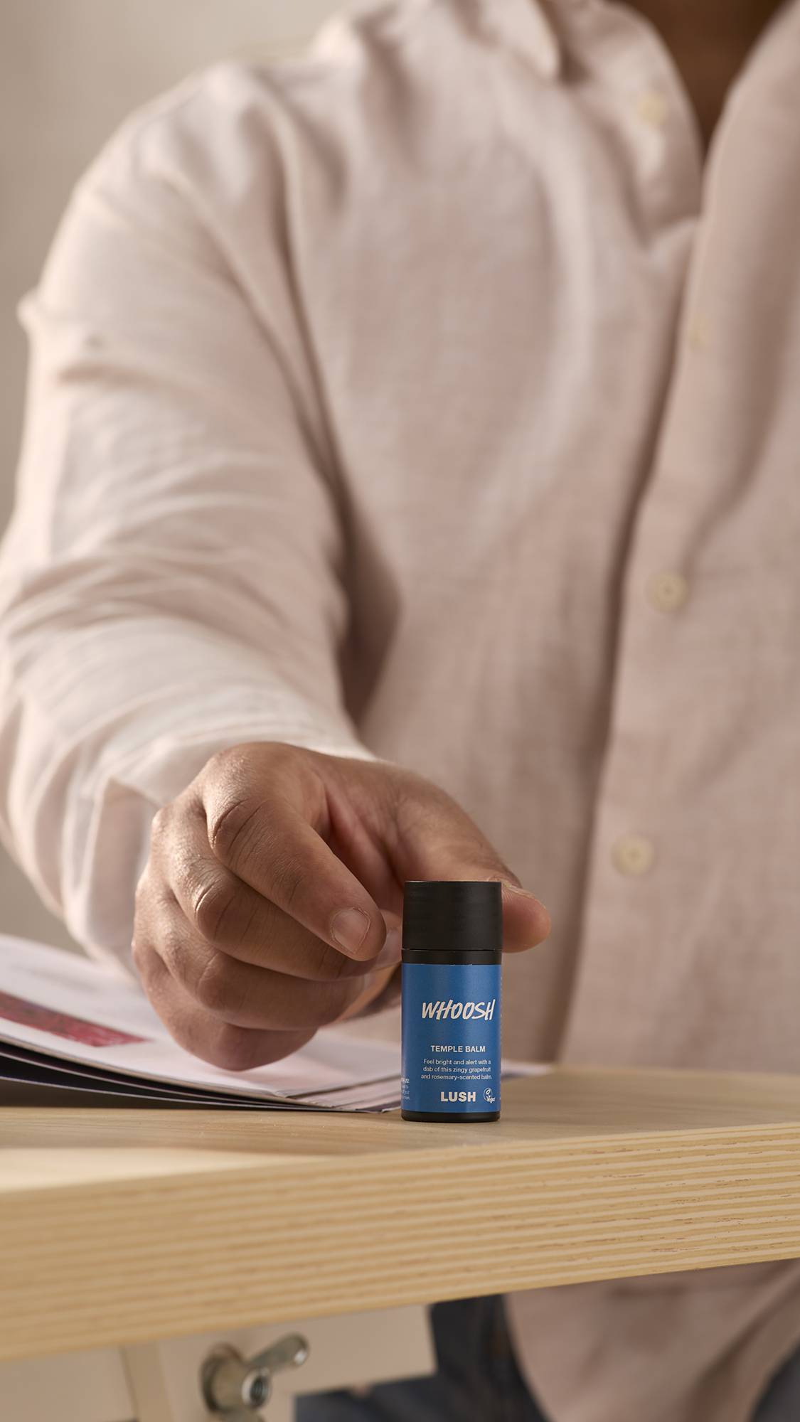 The image shows a close-up of the model sat at a desk in a shirt as they reach for the Whoosh temple balm which is sat on the desk corner facing the camera.