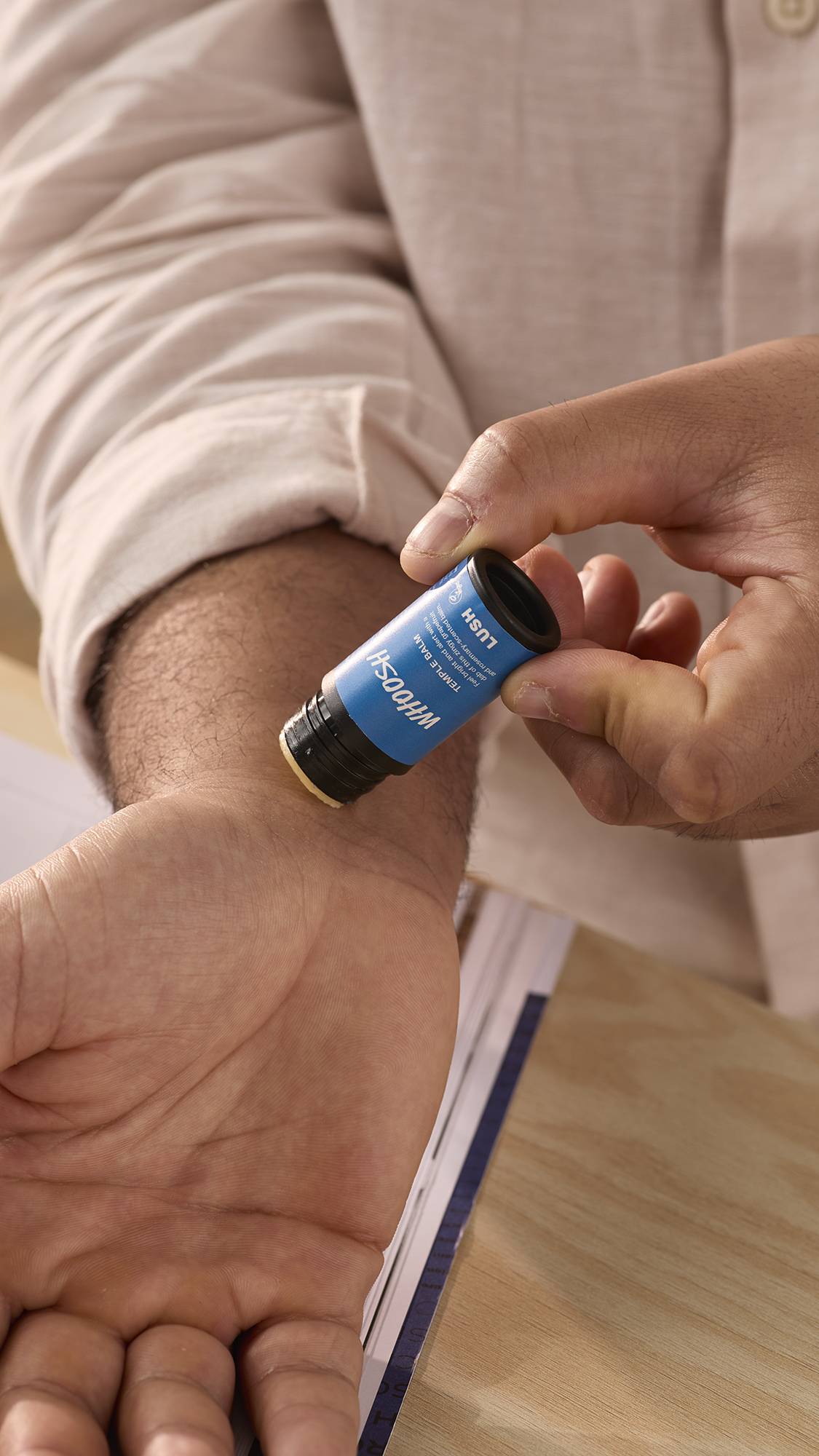 The image shows a close-up of the model's forearms and hands as they gently swipe the balm stick across their wrist.