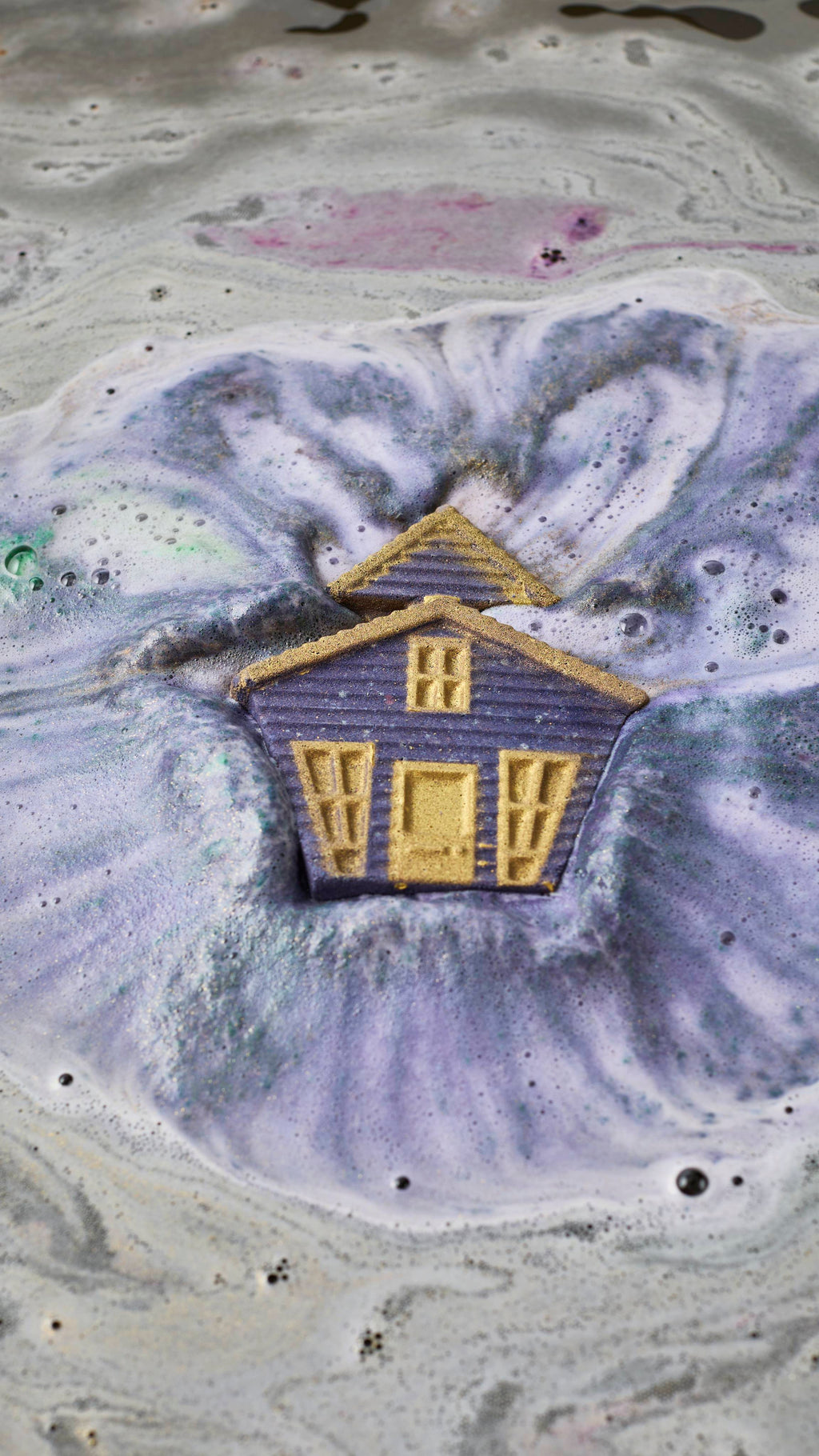 A purple haunted house bath bomb with gold windows and door fizzes and dissolves in murky bathwater, releasing thick swirls of purple and silver-grey foam and bubbles.