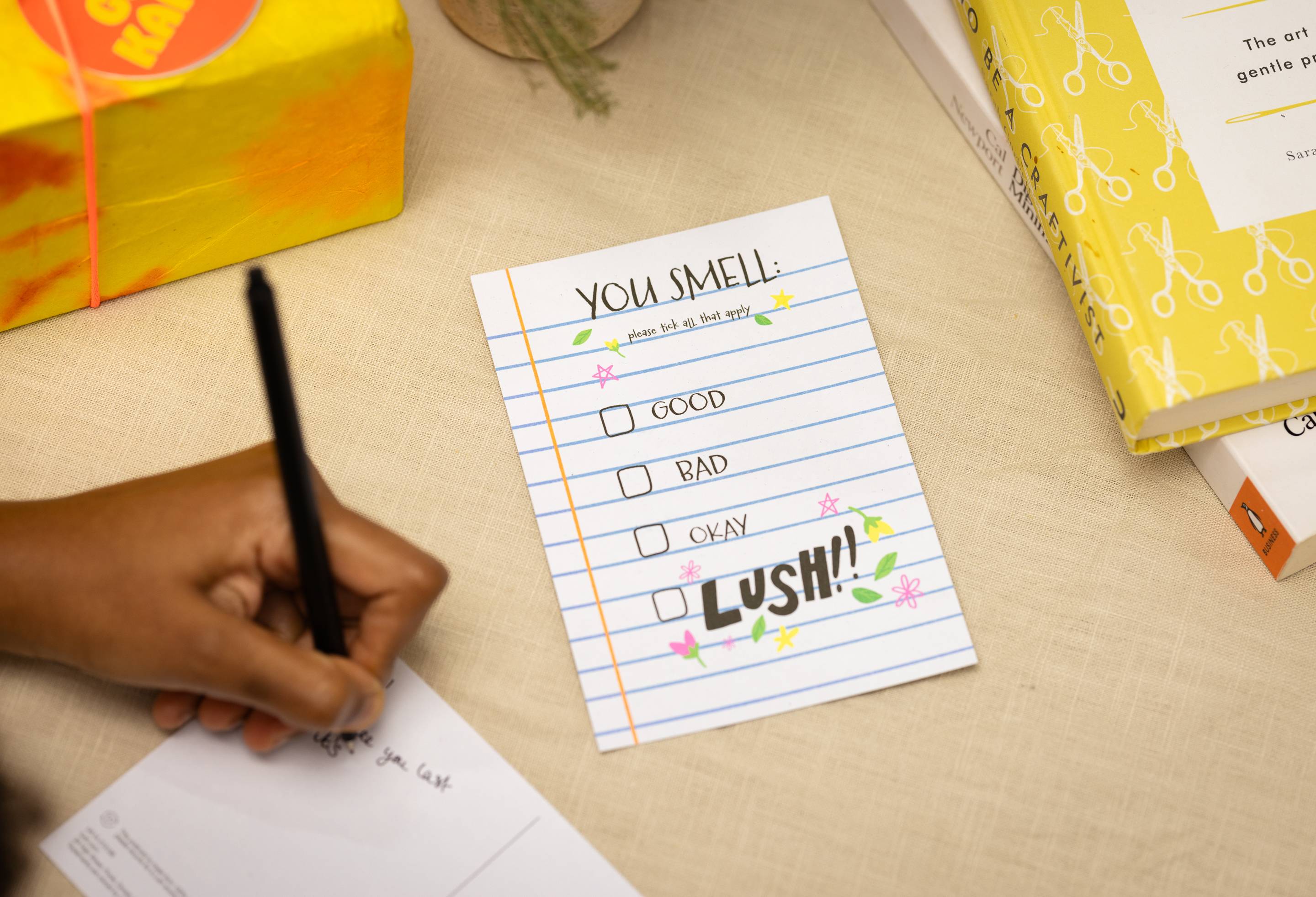 The You Smell: Good, Bad, Okay or LUSH postcard laying on a table as the model writes another card.